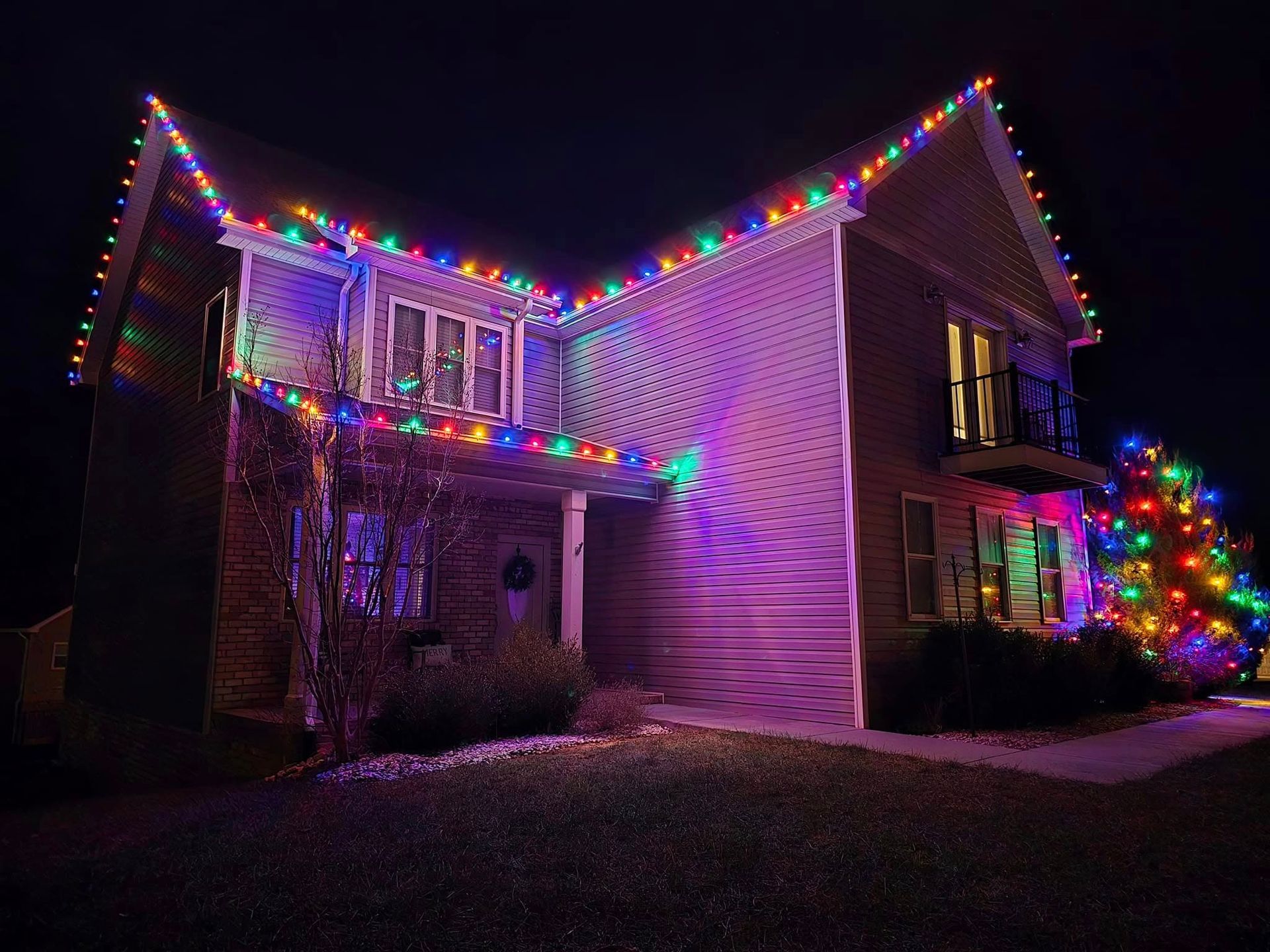 House at night decorated with colorful Christmas lights along the roof and a tree in front.