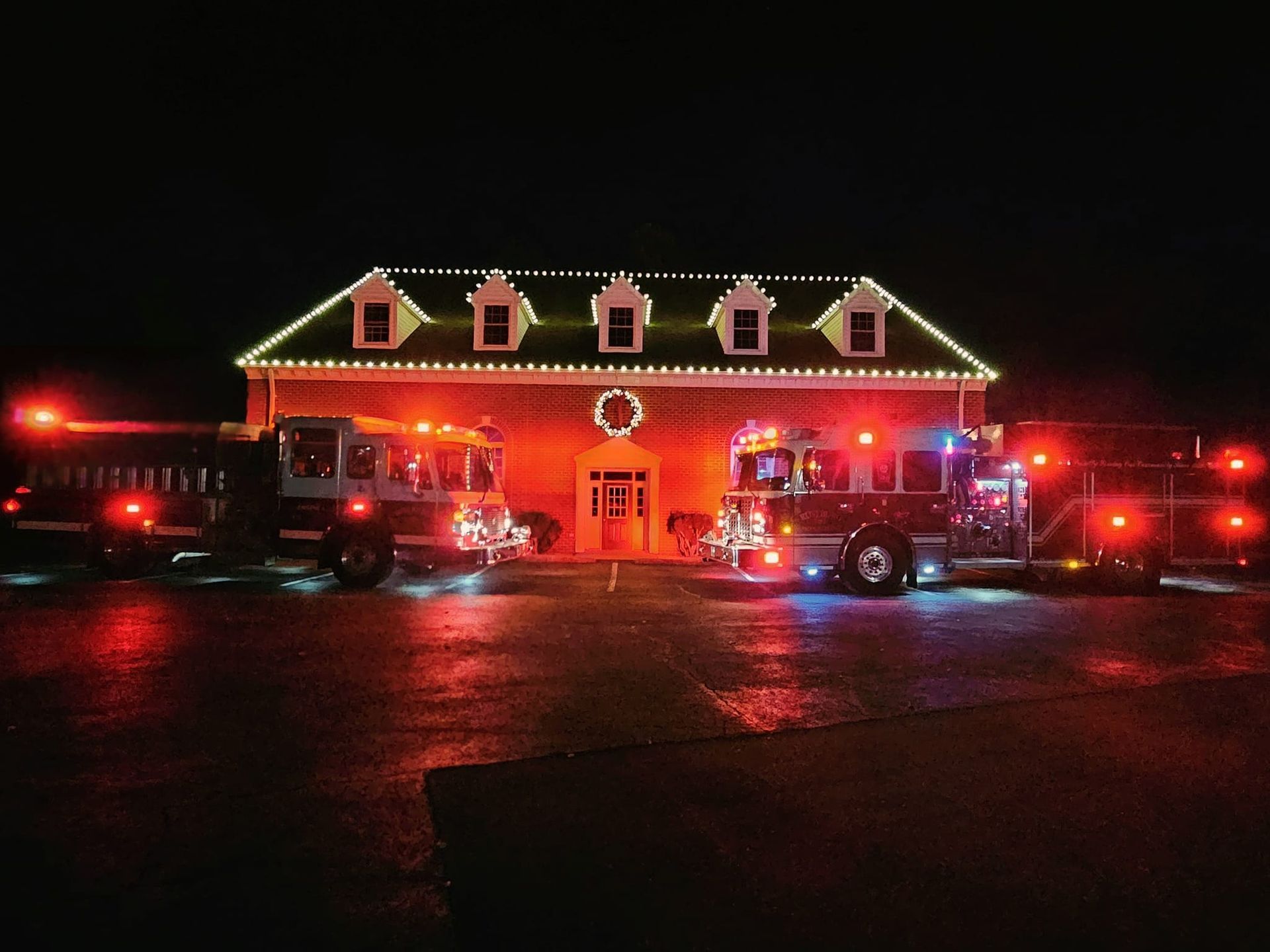 Fire station at night, decorated with Christmas lights, fire trucks with flashing lights parked in front.