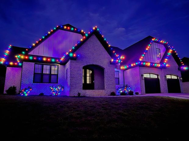 A brick house at night decorated with colorful Christmas lights along the roof and bushes.
