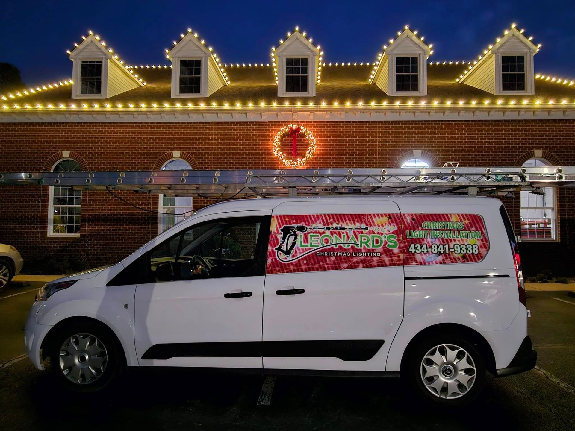 White van with ladder parked in front of a building decorated with Christmas lights at night.