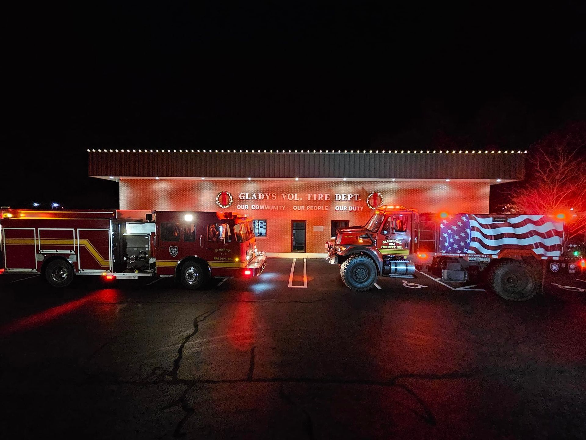 Fire trucks and an American flag-draped casket outside a fire station at night, lit by red and white lights.