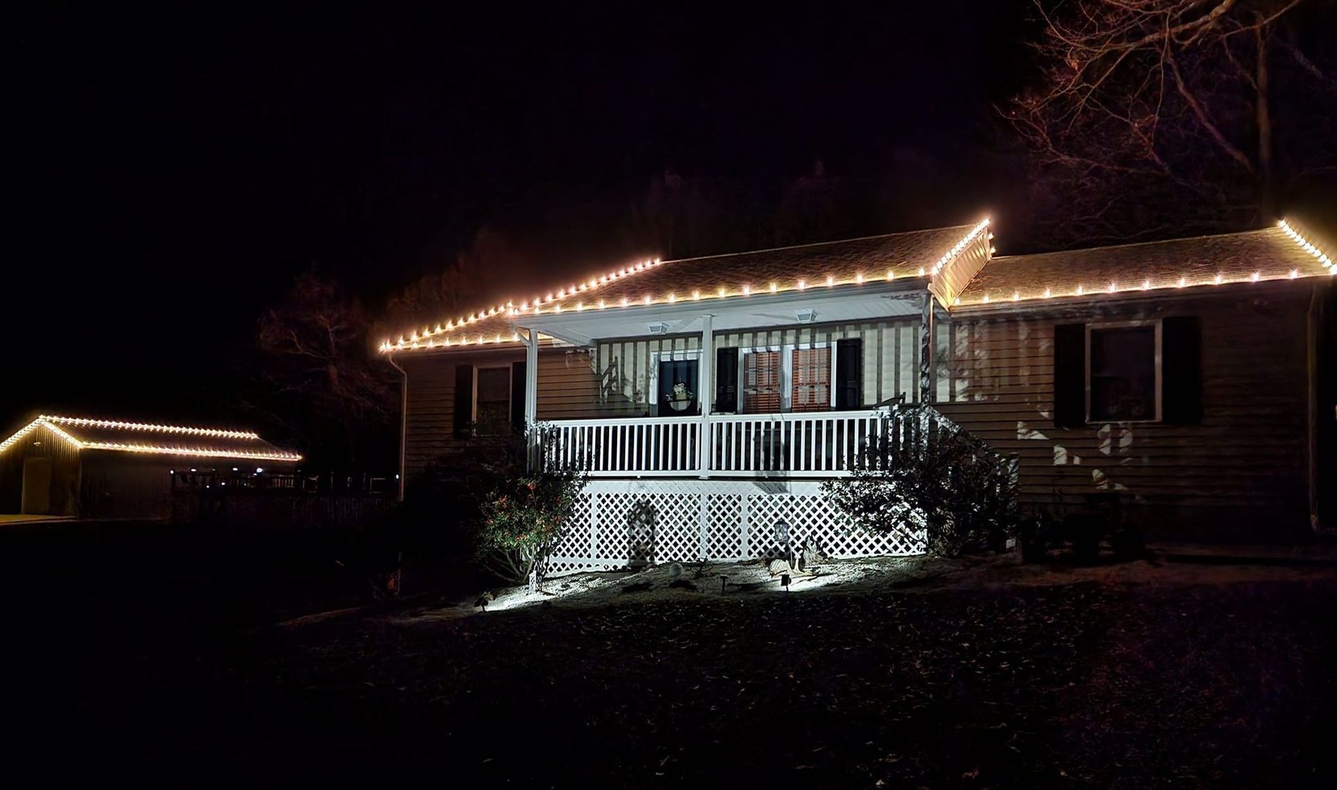 House at night, illuminated with festive lights along the roofline, porch, and a detached structure.