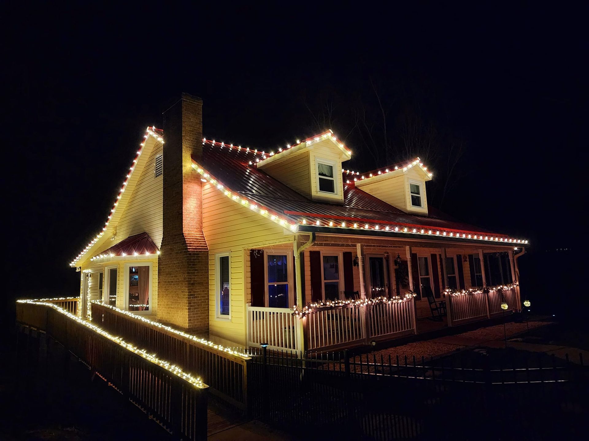 House at night, illuminated with white Christmas lights along the roof and fence, with a porch and windows.