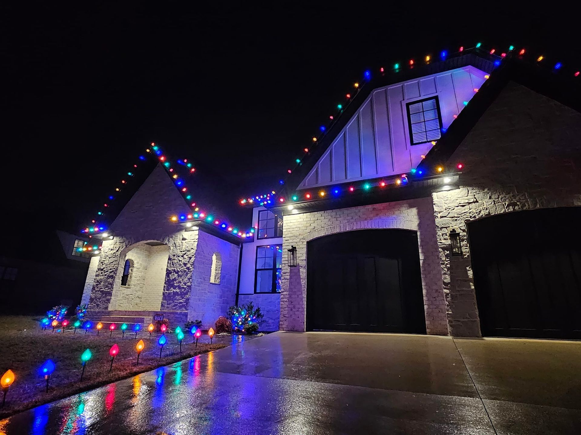 House decorated with colorful Christmas lights at night, reflecting on wet pavement.