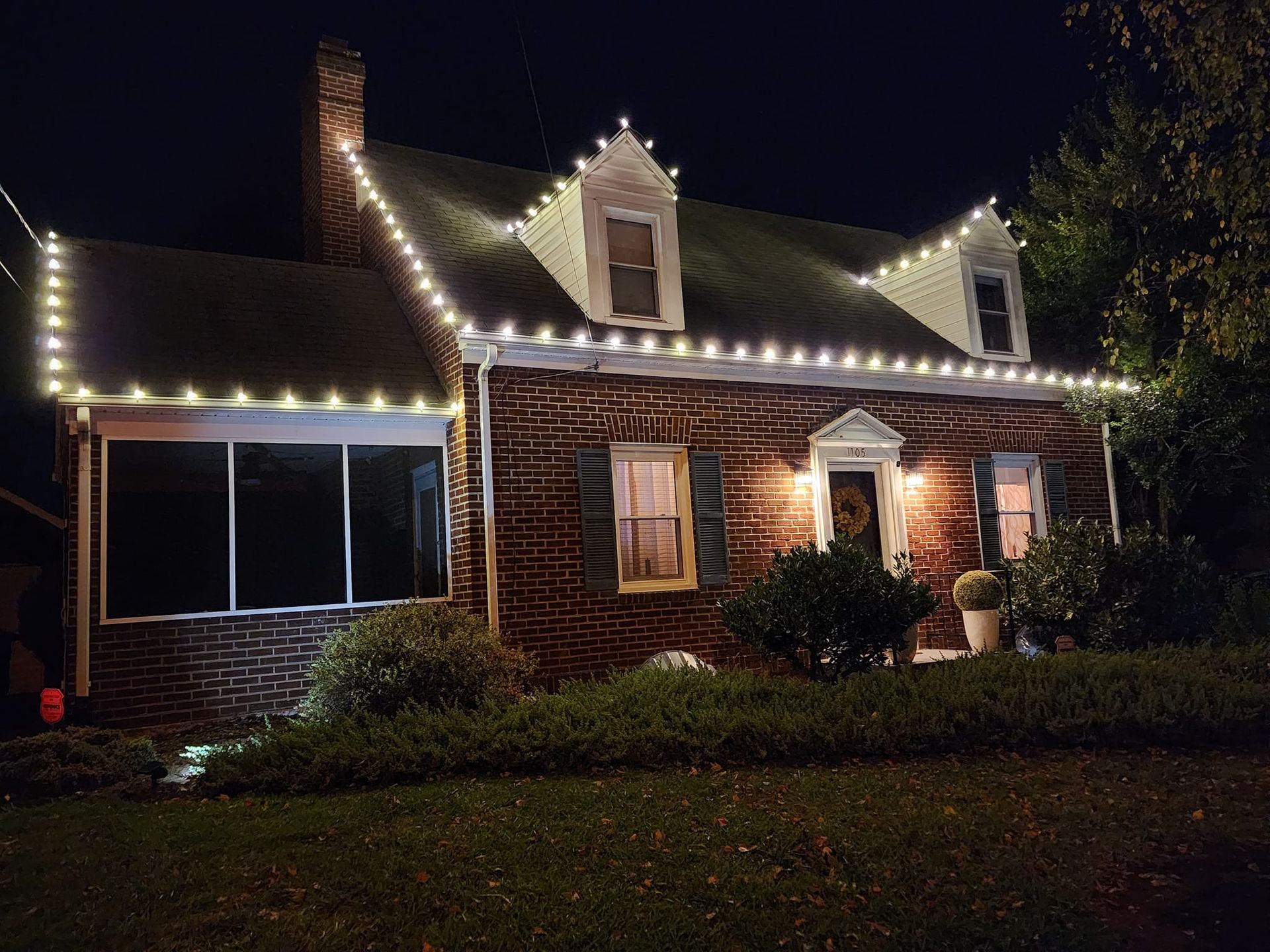 Brick house at night, decorated with white Christmas lights along roofline and windows.
