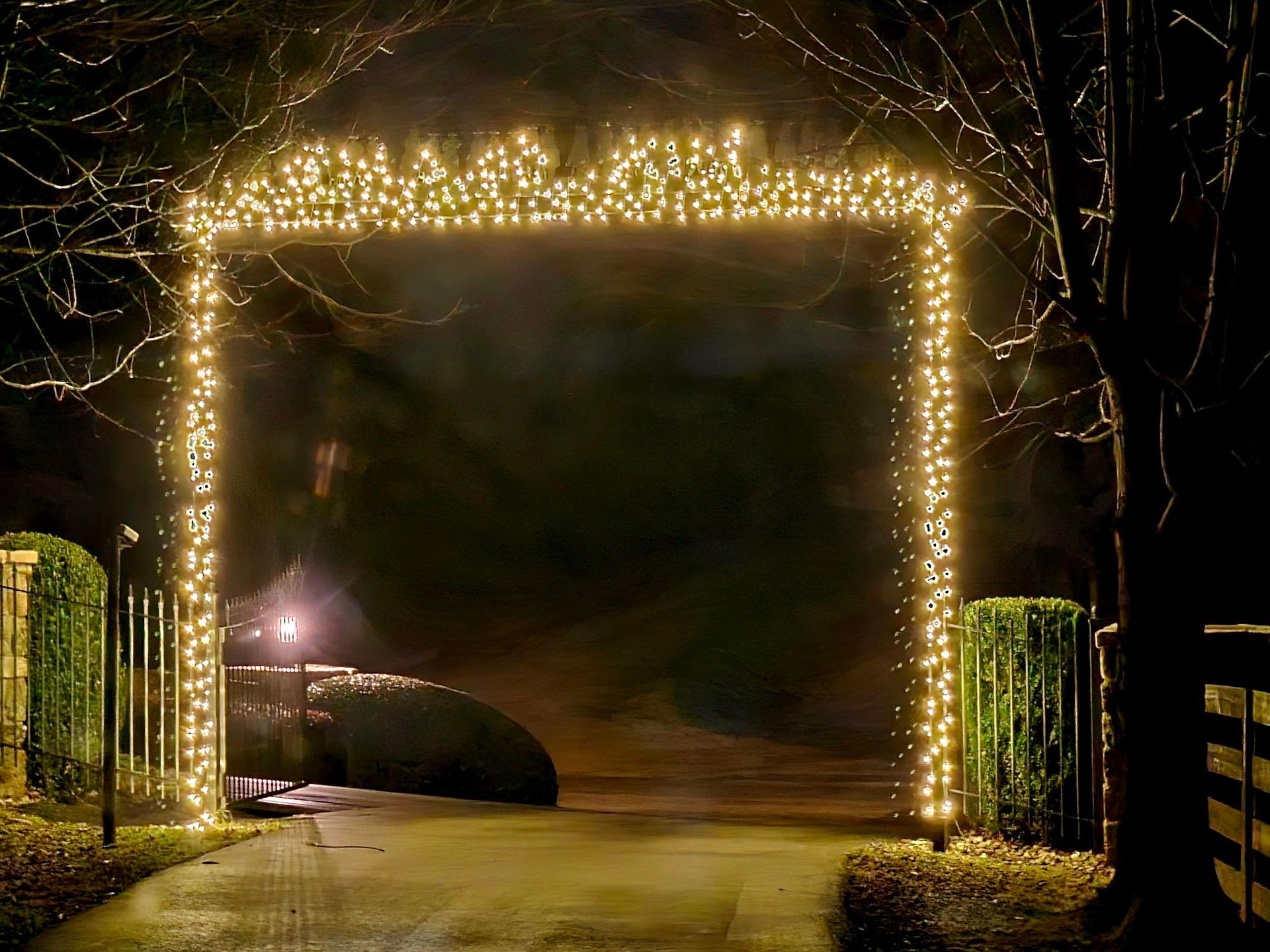 An archway outlined with golden Christmas lights illuminates a dark driveway and surrounding trees.