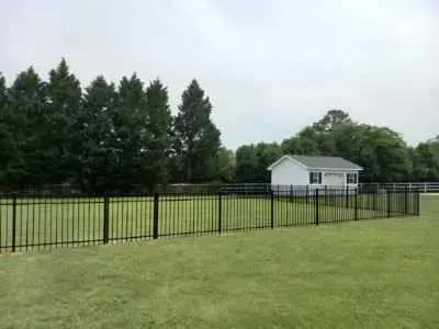 A black fence surrounds a lush green field with a white house in the background.