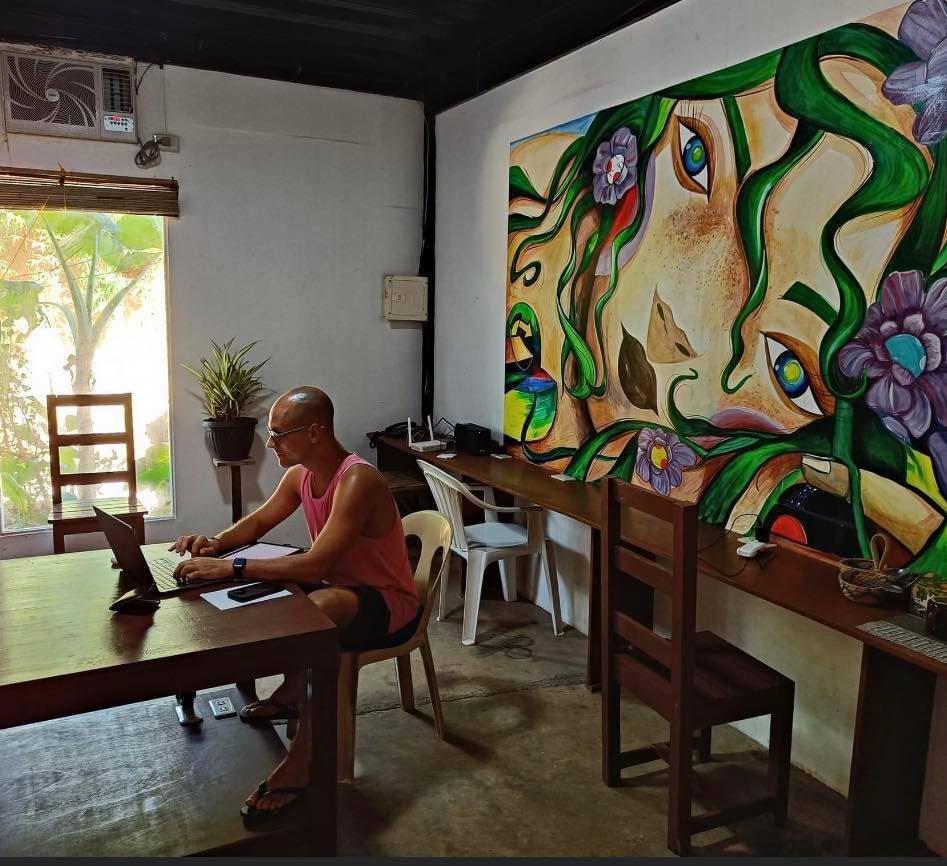A man sits at a table with a laptop in front of a painting on the wall
