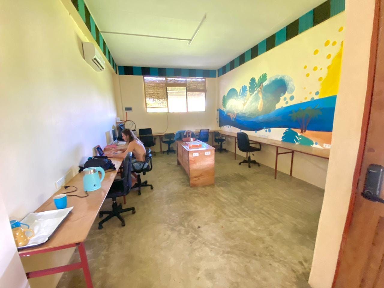 A group of people are sitting at desks in a room with a mural on the wall.