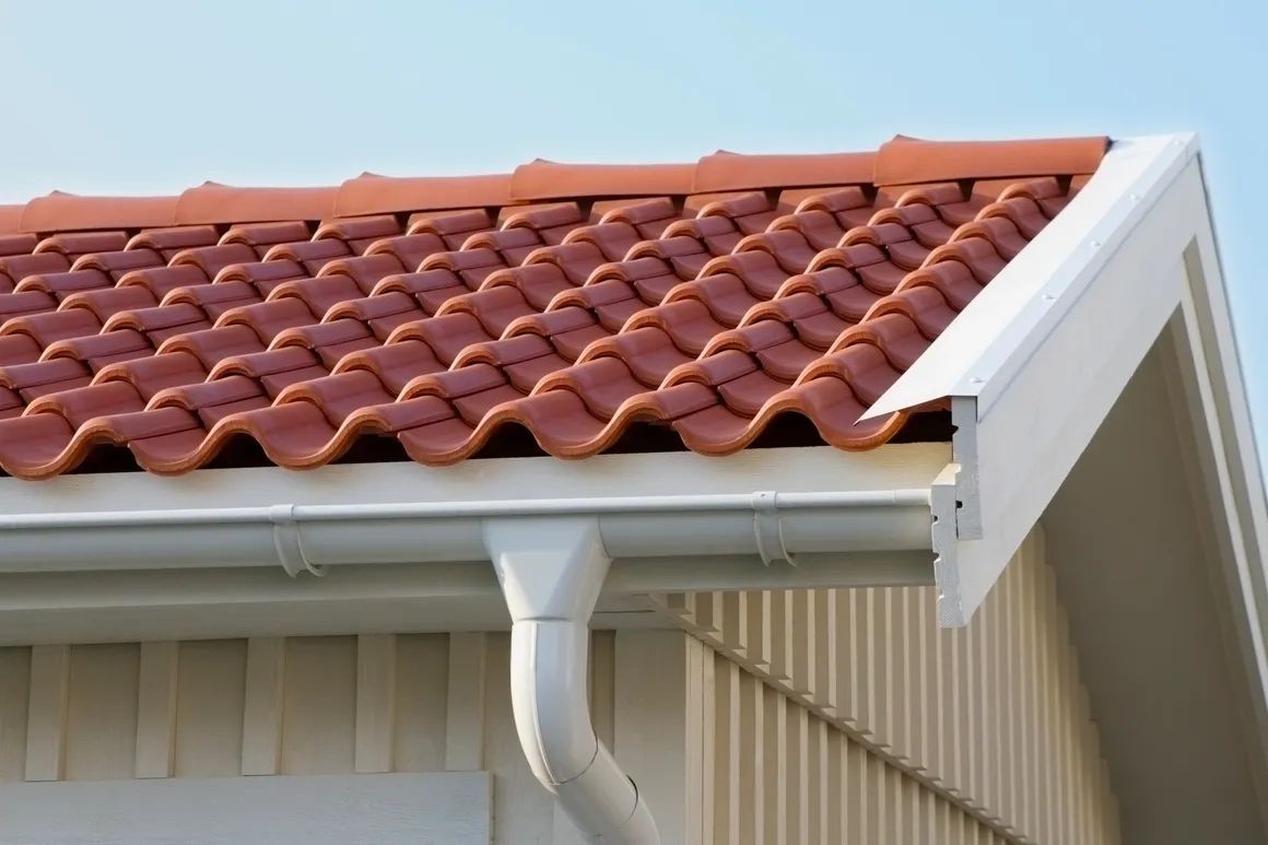 Close-up of a house roof with red tiles, white trim, and a rain gutter against a beige exterior