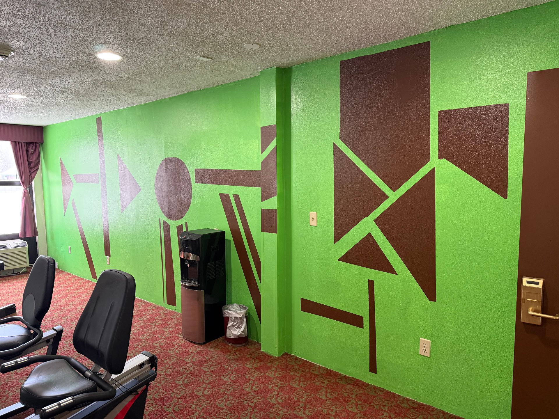 Green and brown geometric mural on wall, two exercise bikes, water cooler.