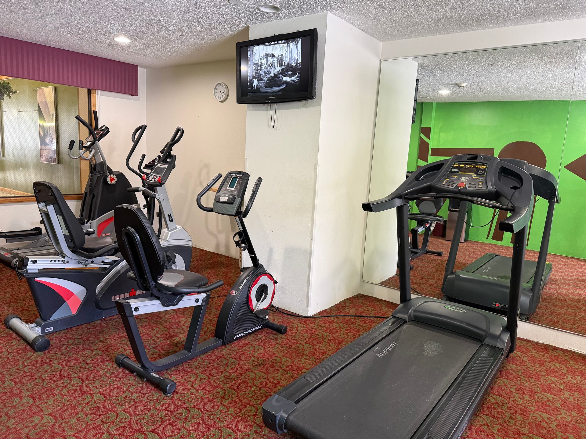 Gym with recumbent bikes, elliptical, stationary bike, treadmill, television, and mirror on red carpet.