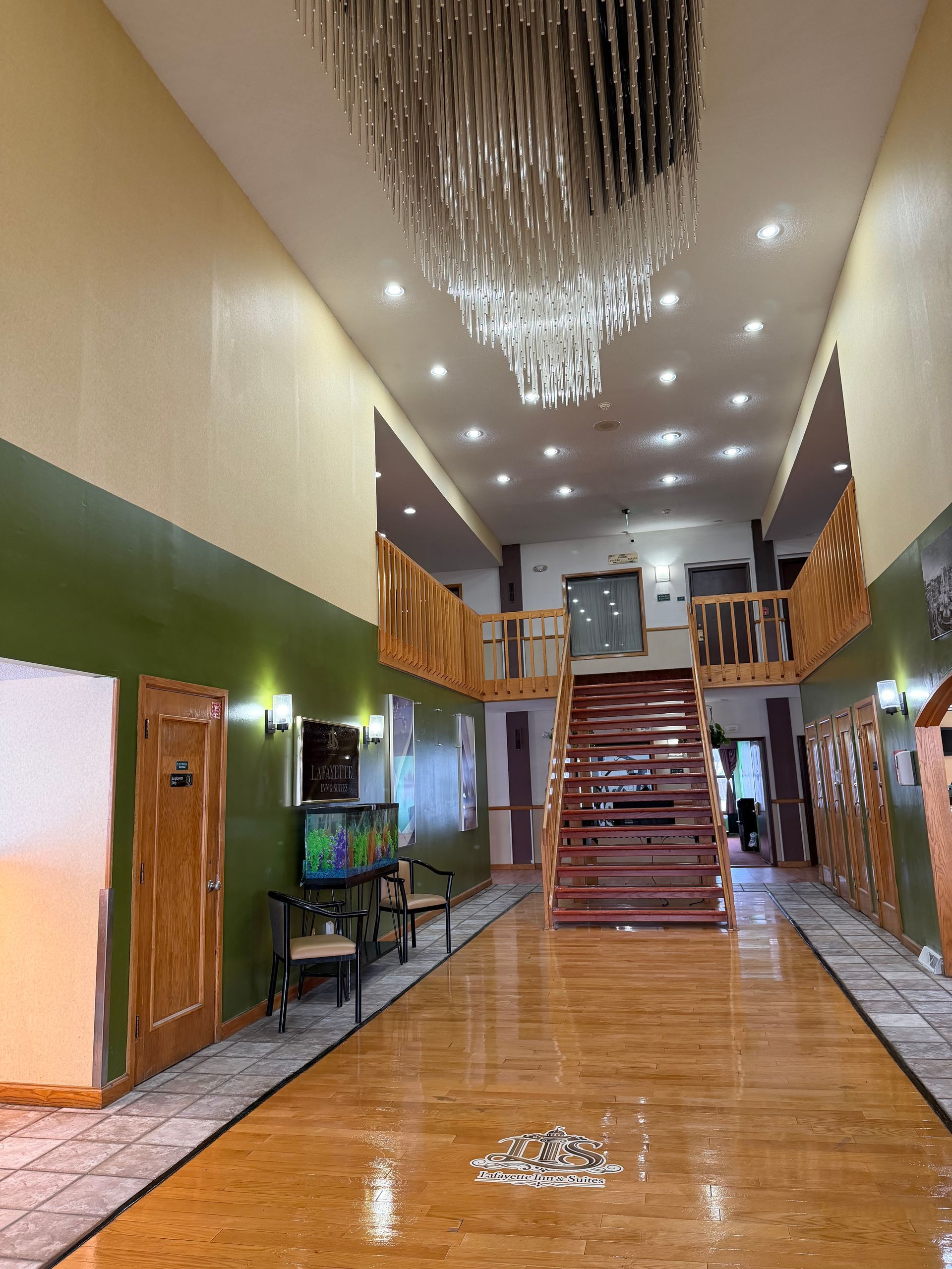 Long hallway with chandelier, wooden staircase, and second-floor balcony. Green and yellow walls with wooden flooring.