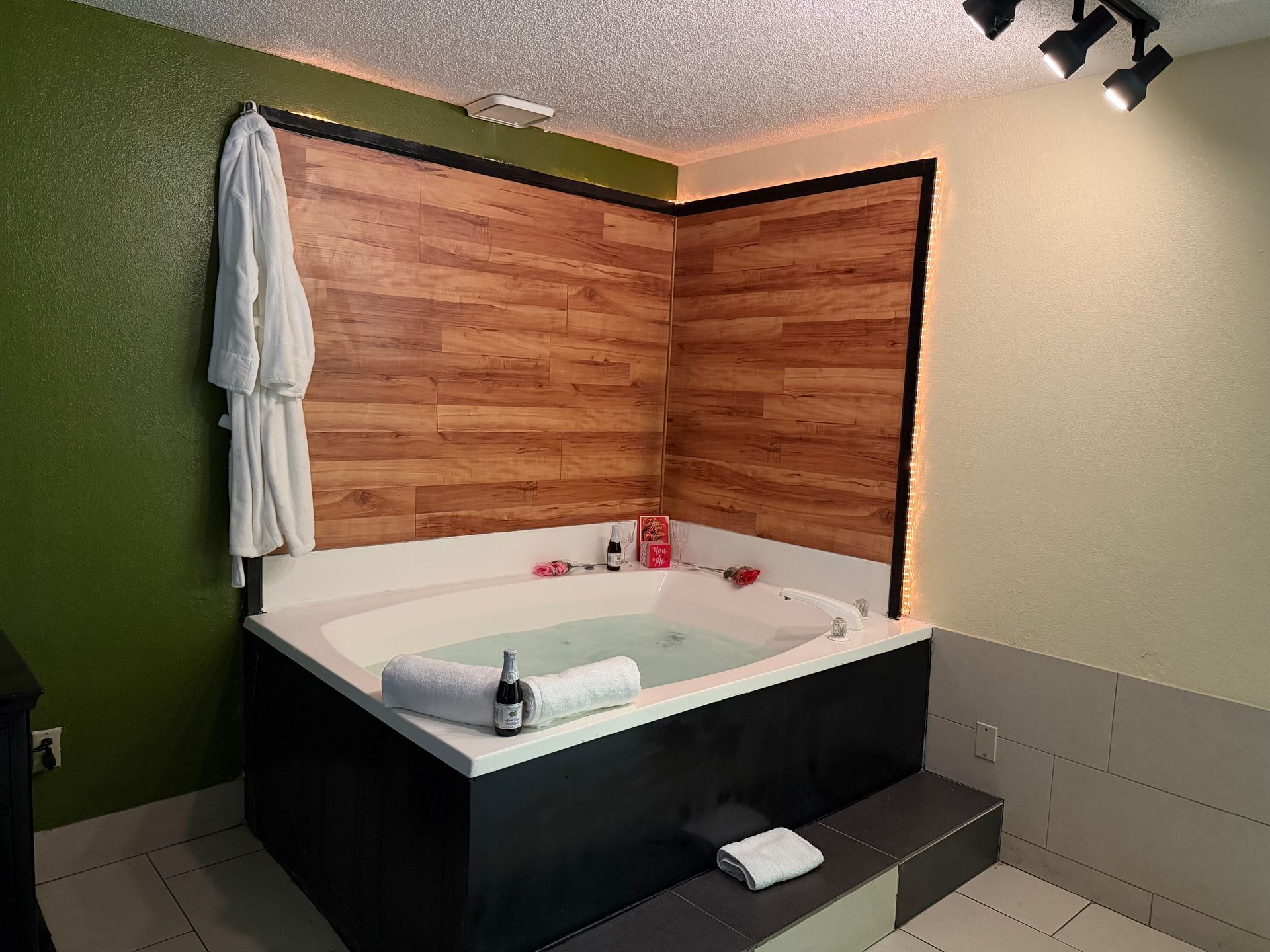 Hot tub in a room with a wood-paneled wall and robe hanging, towels, and lights.