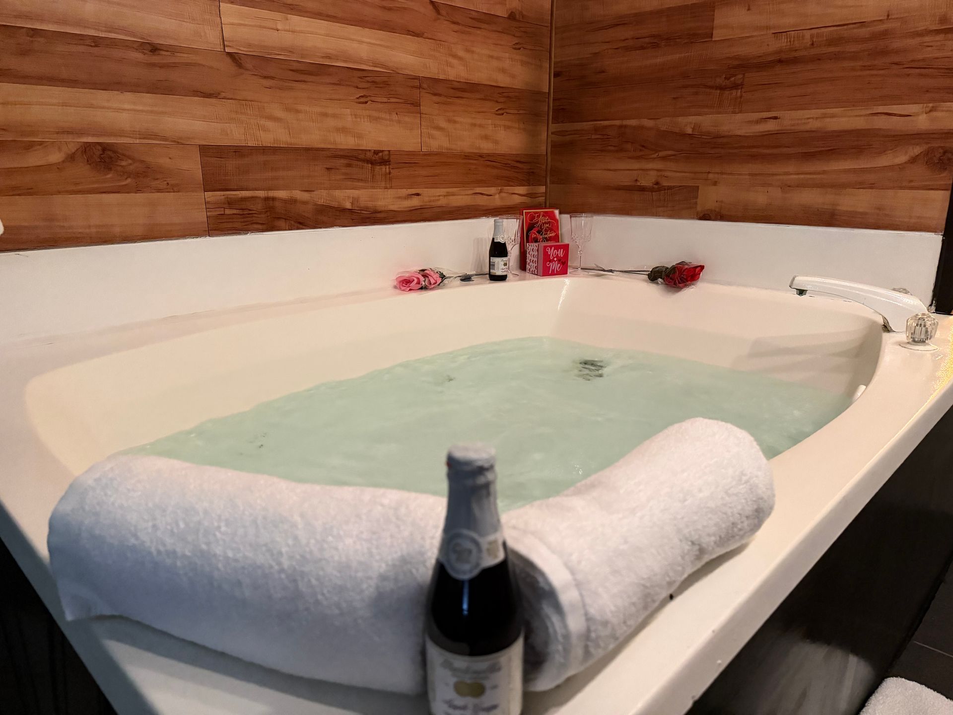 Hot tub filled with water, rolled towels, wine bottle, and rose on the side. Wooden wall in the background.
