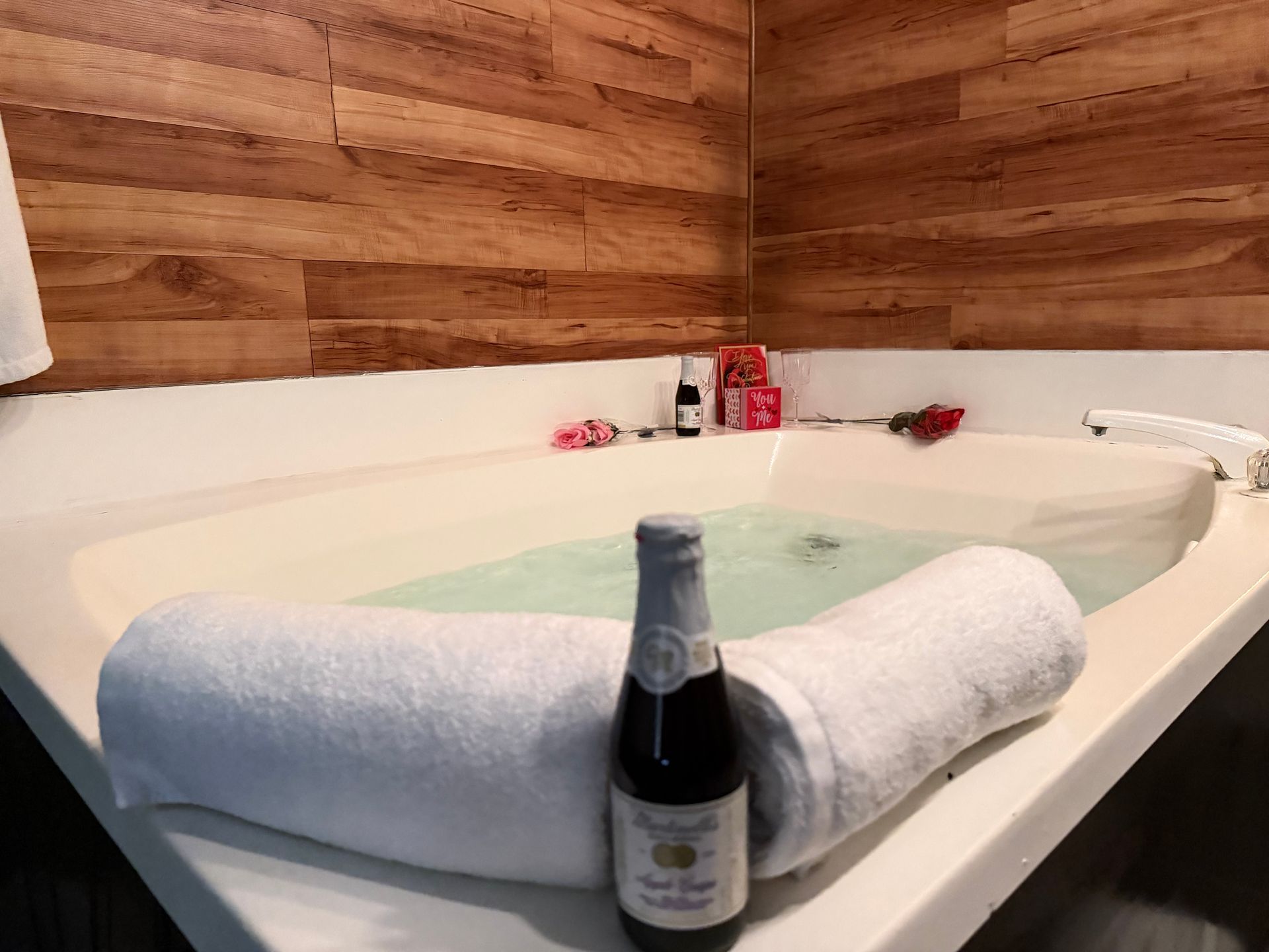 A jacuzzi tub with towels, champagne, and rose petals, set in a room with wood paneling.