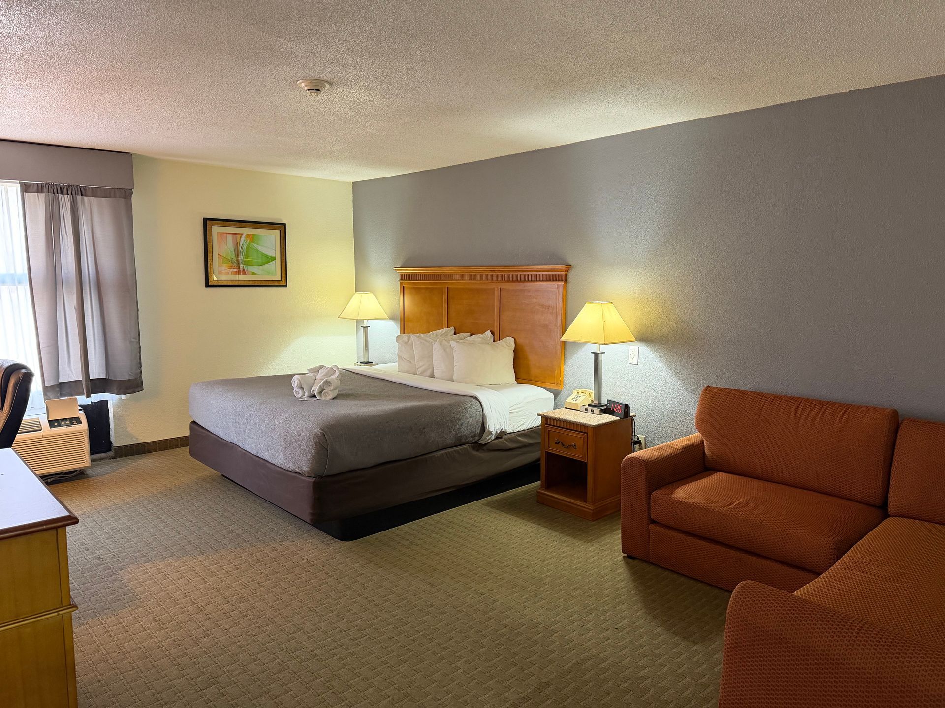 Hotel room with king bed, sofa, desk, and bedside lamps. Beige walls and carpet.