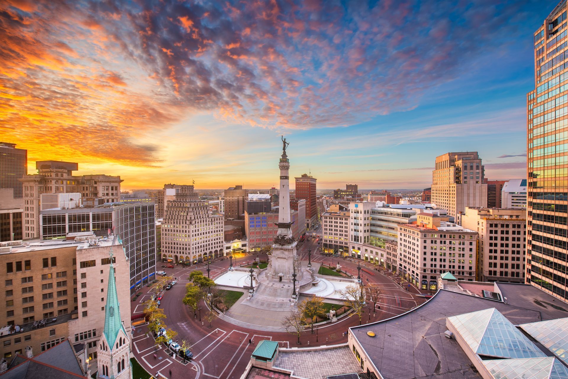 Indianapolis city skyline at sunset, Monument Circle prominent, with buildings and colorful sky.