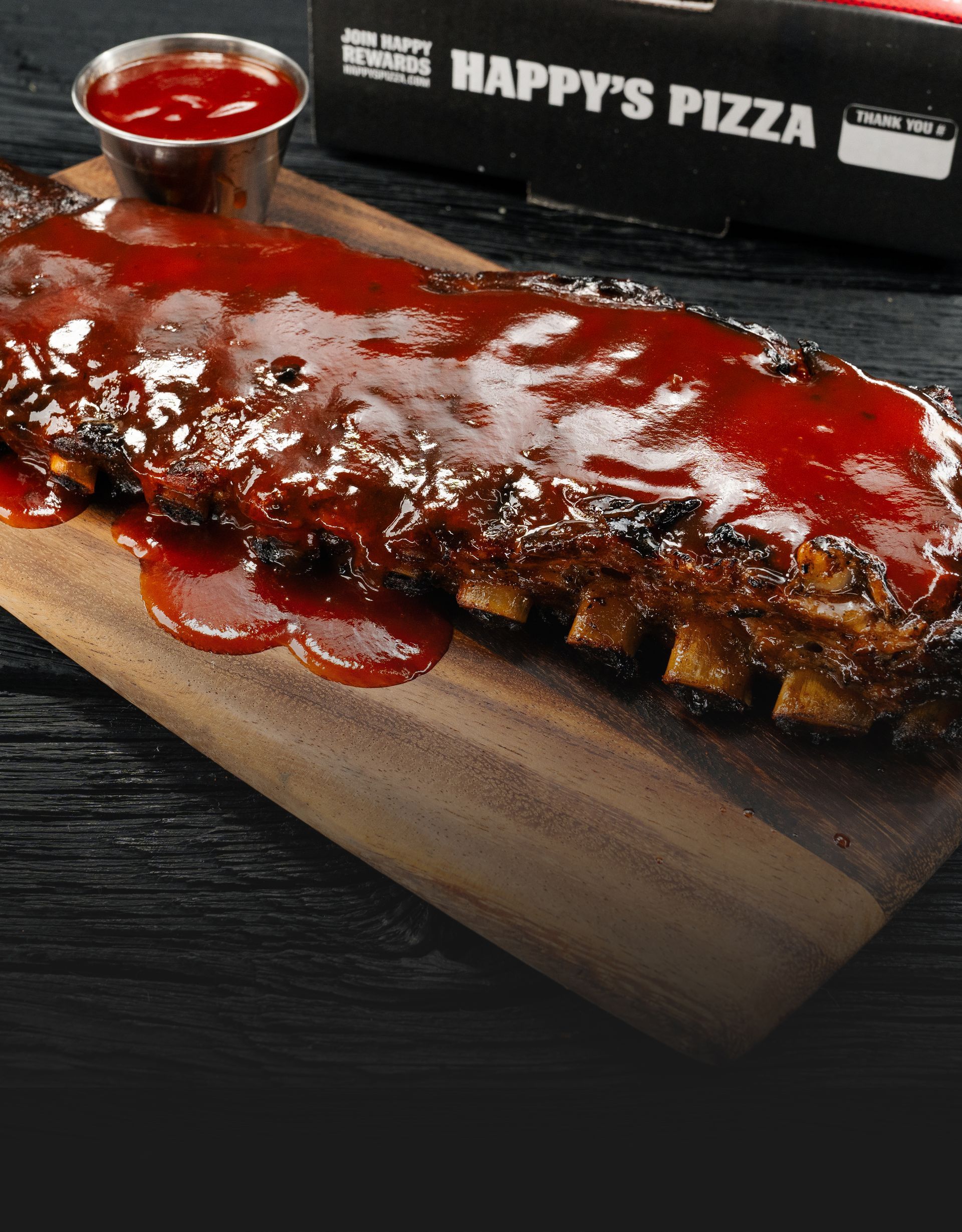 A full slab of BBQ ribs glazed with sauce, served on a wooden cutting board with a cup of BBQ Sauce