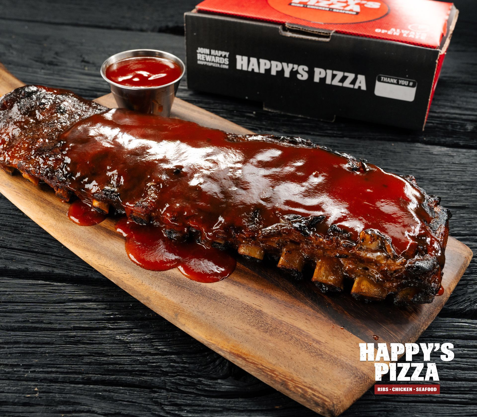 A very long piece of meat is on a wooden cutting board next to a happy 's pizza box.