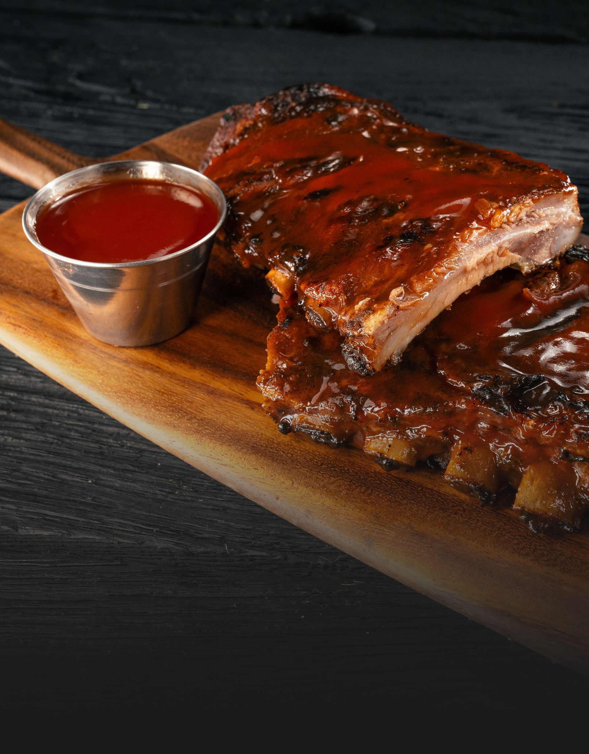 A wooden cutting board topped with ribs and a cup of sauce.