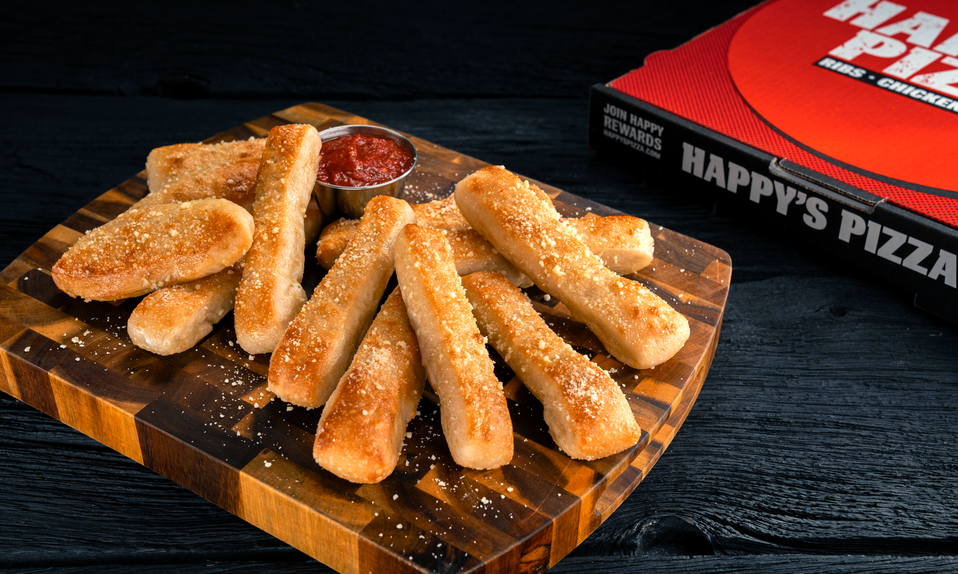 A wooden cutting board topped with cheese sticks next to a box of happy 's pizza.