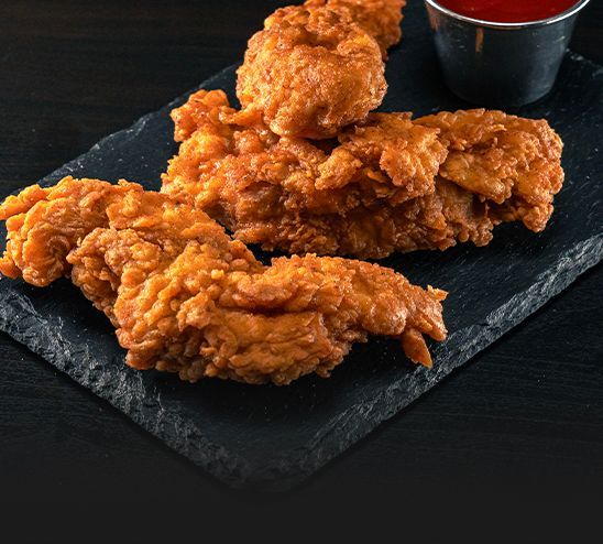 Fried chicken is sitting on a slate cutting board next to a cup of sauce.