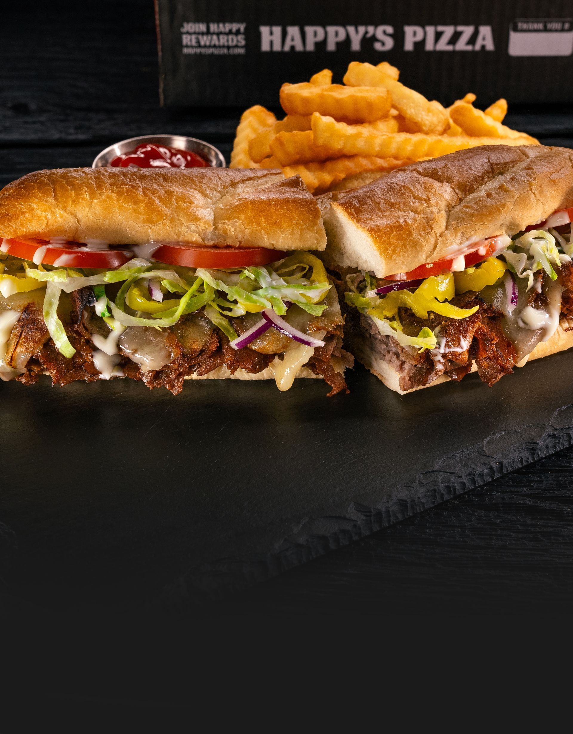 A sub sandwich with lettuce tomatoes and pickles next to a happy 's pizza box