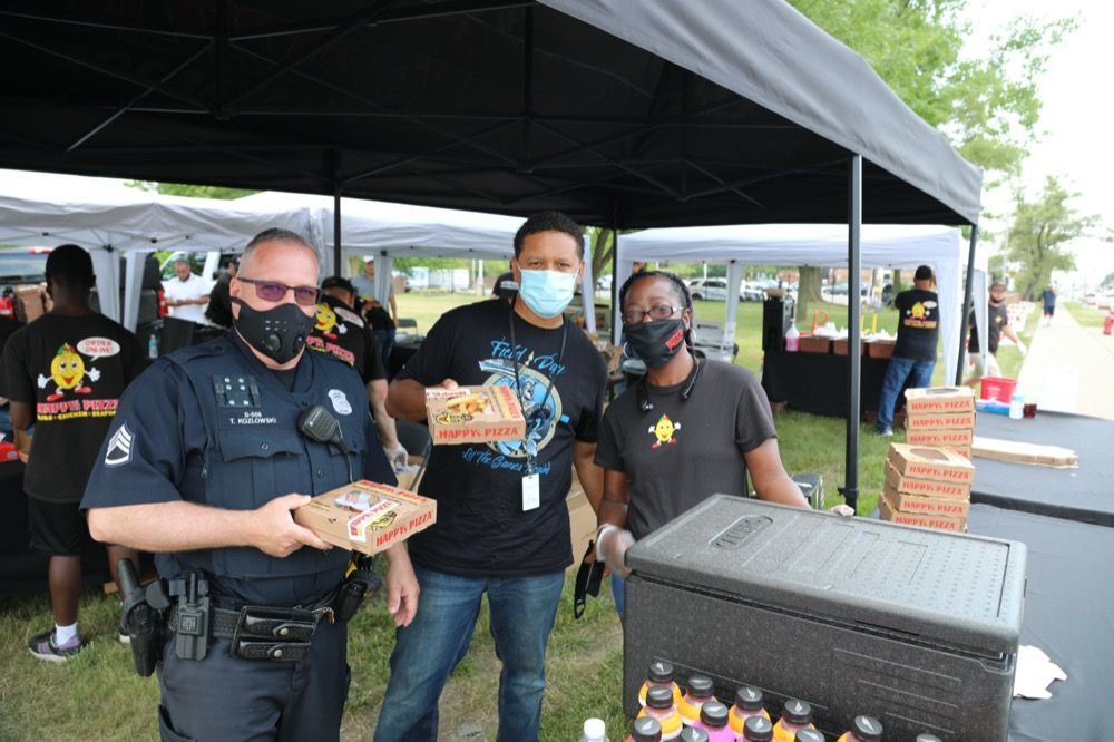A group of people wearing masks are standing under a tent holding pizzas.