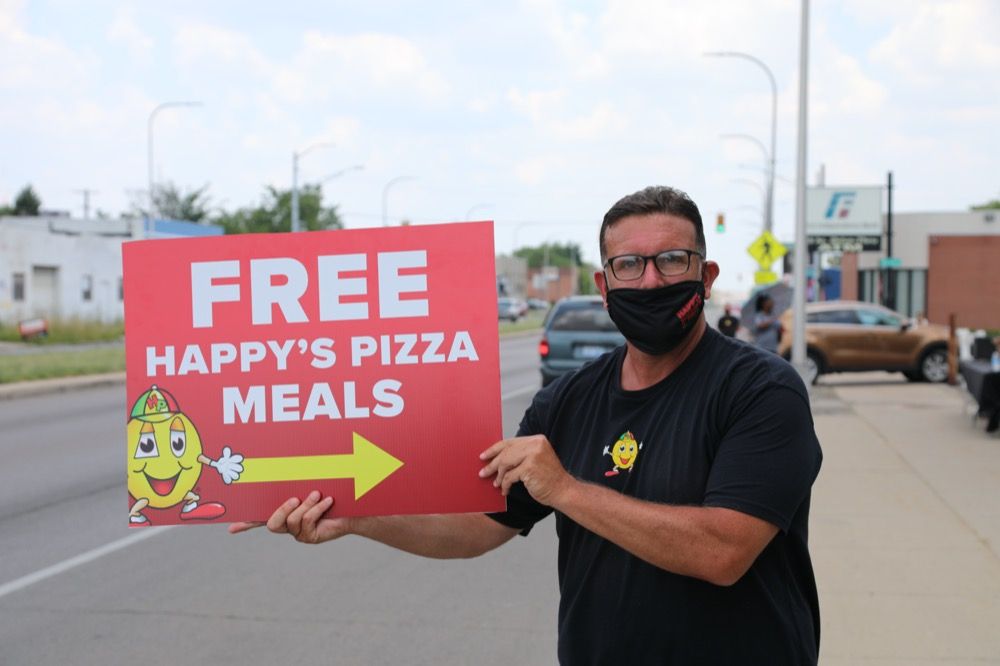 A man wearing a mask is holding a sign that says free happy 's pizza meals.