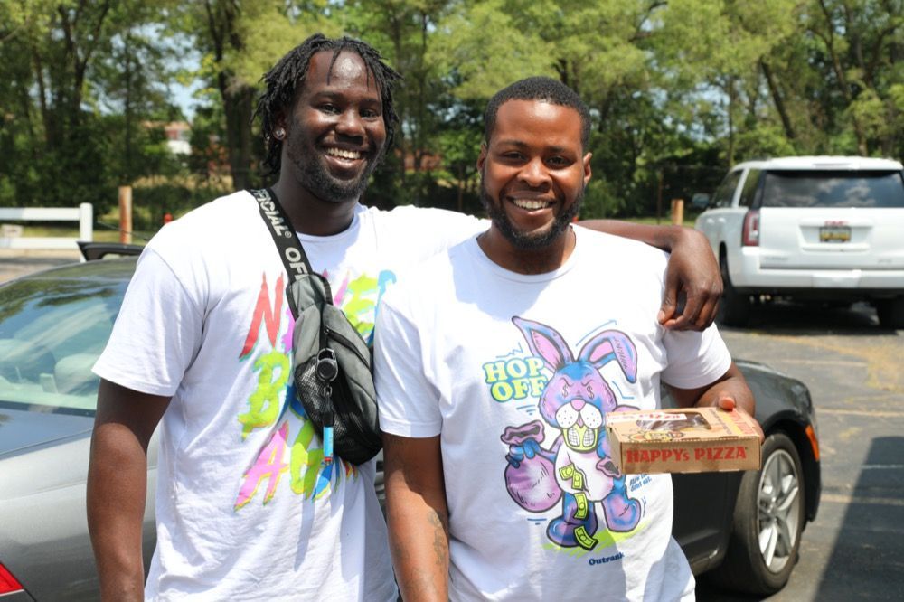 Two men are standing next to each other in a parking lot holding a box of pizza.