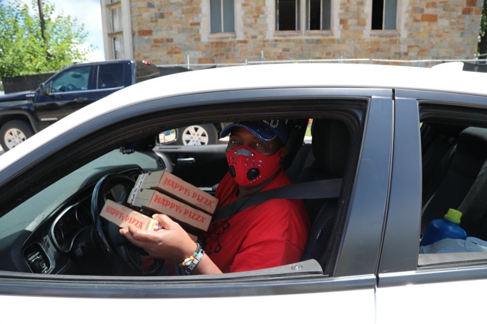 A man wearing a mask is sitting in a car holding two boxes of pizza.