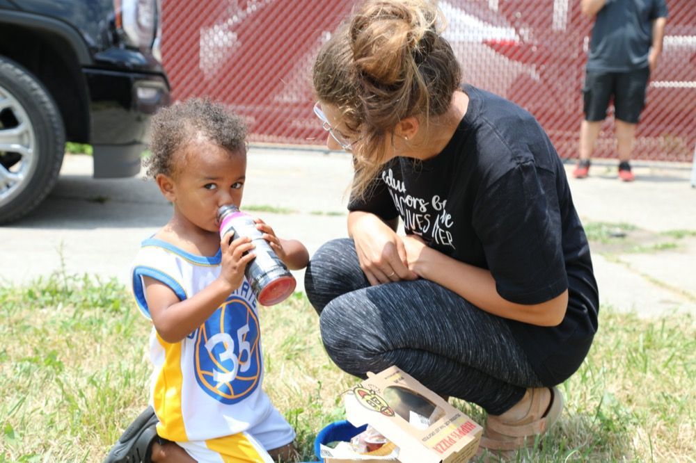 A woman is kneeling down next to a little boy drinking from a bottle.