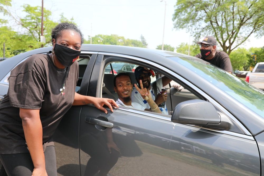 A woman wearing a mask is standing next to a car with people in it.