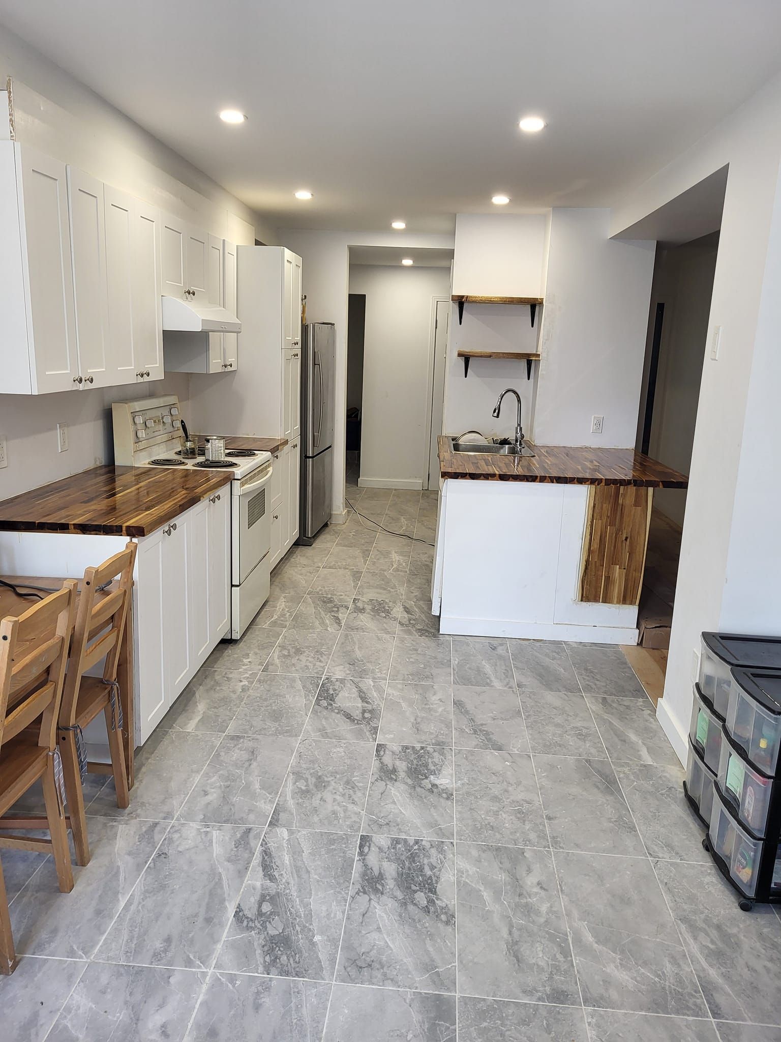 Une cuisine longue et étroite avec des armoires blanches, des plans de travail en bois et un sol en carrelage gris. Elle comprend une cuisinière, un réfrigérateur et un îlot central avec évier.