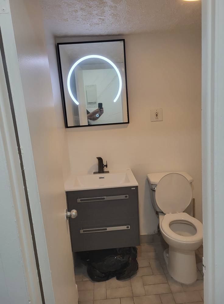 Une petite salle de bain avec un meuble-lavabo gris, des toilettes et un miroir avec un halo lumineux. Un sac poubelle noir est posé par terre.