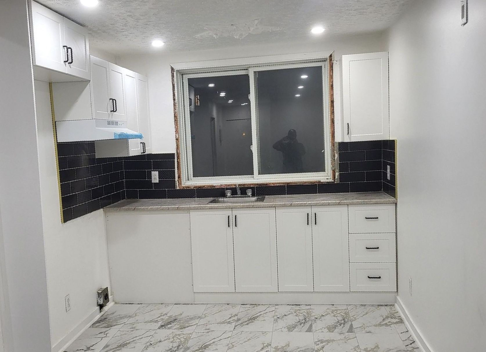Une petite cuisine moderne avec des armoires blanches, une crédence en carrelage noir et une fenêtre. Le plan de travail est gris clair et le sol est marbré.