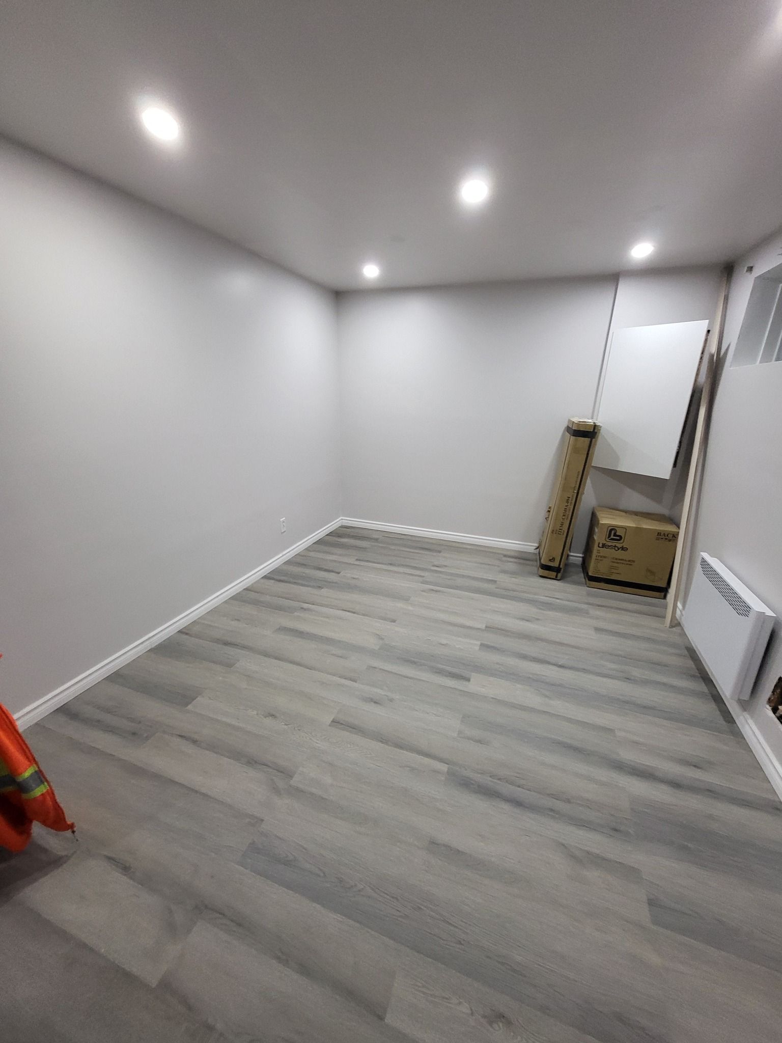 Pièce vide avec parquet gris, murs blancs et éclairage encastré. Quelques objets sont rangés le long du mur du fond.