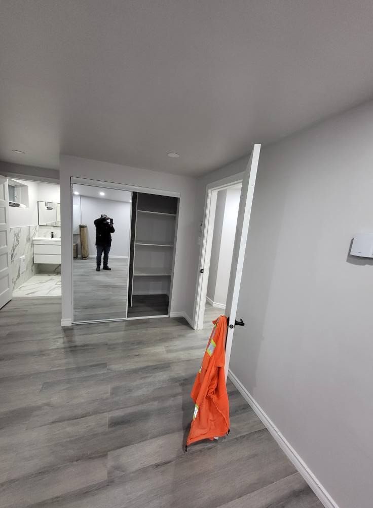Une personne se tient dans une pièce aux murs et au sol gris clair. Il y a un placard avec des étagères et une porte ouverte. Un gilet de sécurité orange est appuyé contre l'encadrement de la porte.