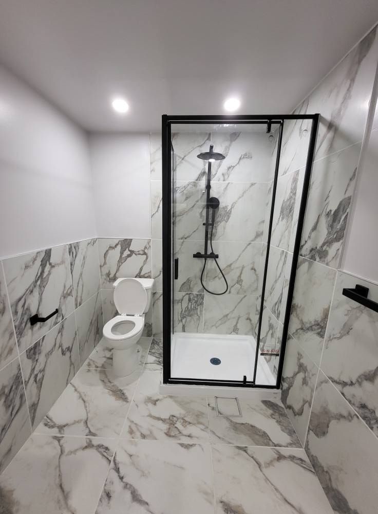 Salle de bain moderne avec carrelage à motifs marbrés, douche à cadre noir, toilettes et robinetterie noire. Murs blancs et éclairage encastré.