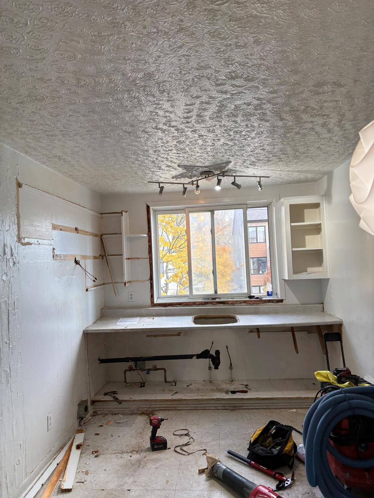 Intérieur d'une pièce en rénovation, avec des montants apparents, un bureau intégré, une fenêtre et des outils. Le plafond présente une finition texturée.