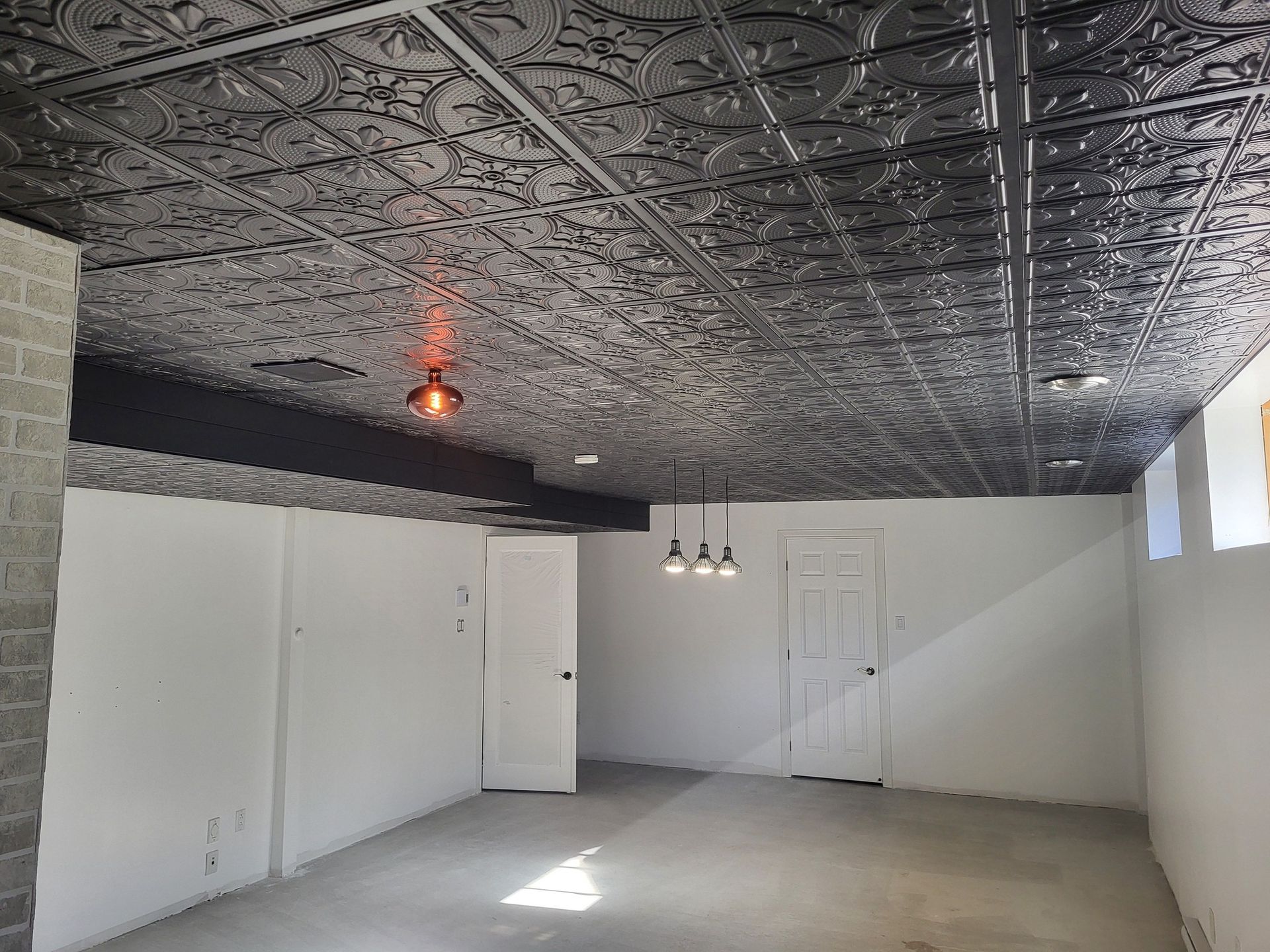 Sous-sol avec plafond en tôle argentée, murs blancs et sol en ciment. Deux portes et un luminaire suspendu sont visibles.