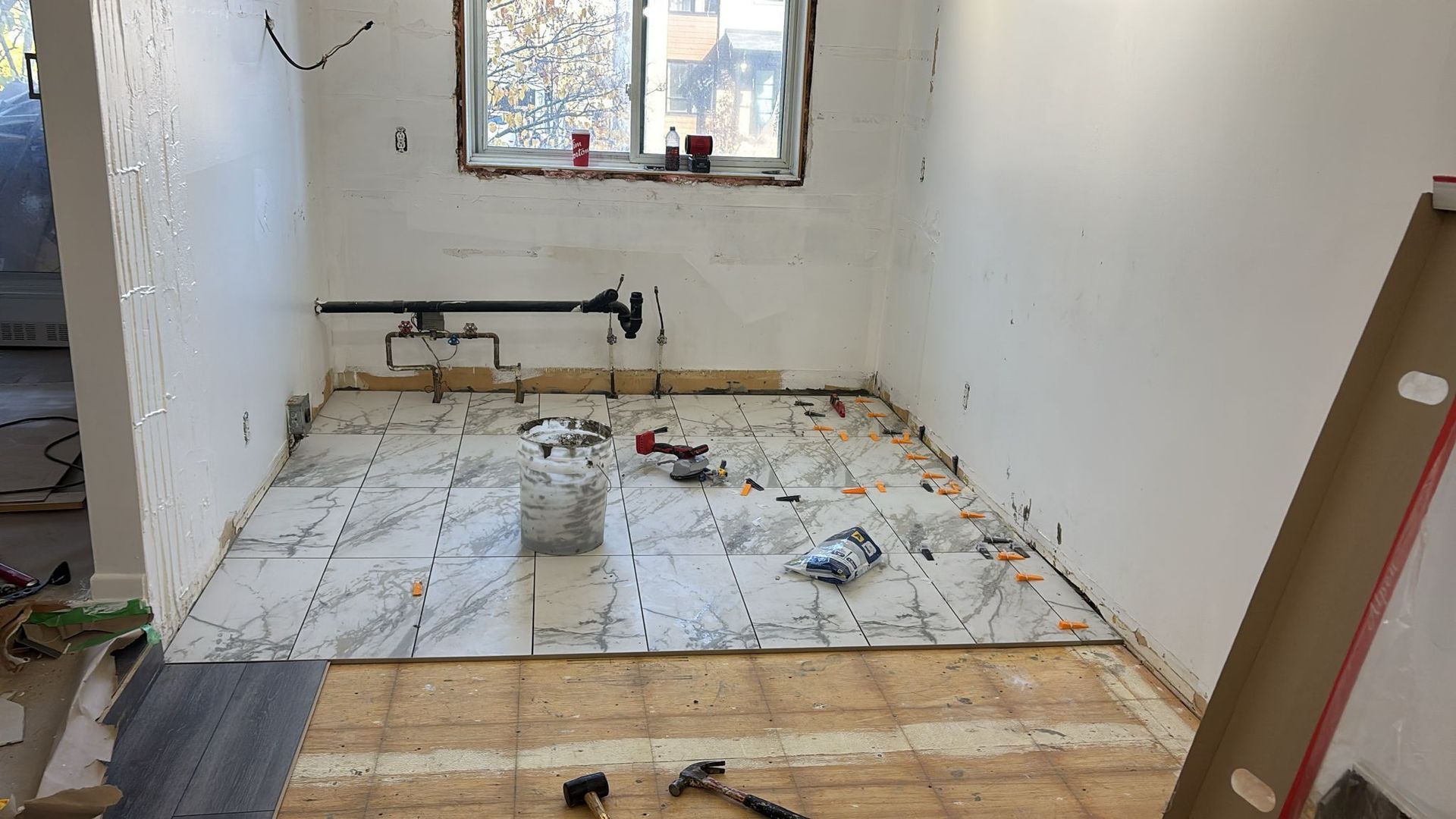 Une pièce en rénovation avec un sol partiellement carrelé, des canalisations apparentes et une fenêtre. Des outils et des matériaux de construction sont éparpillés un peu partout.