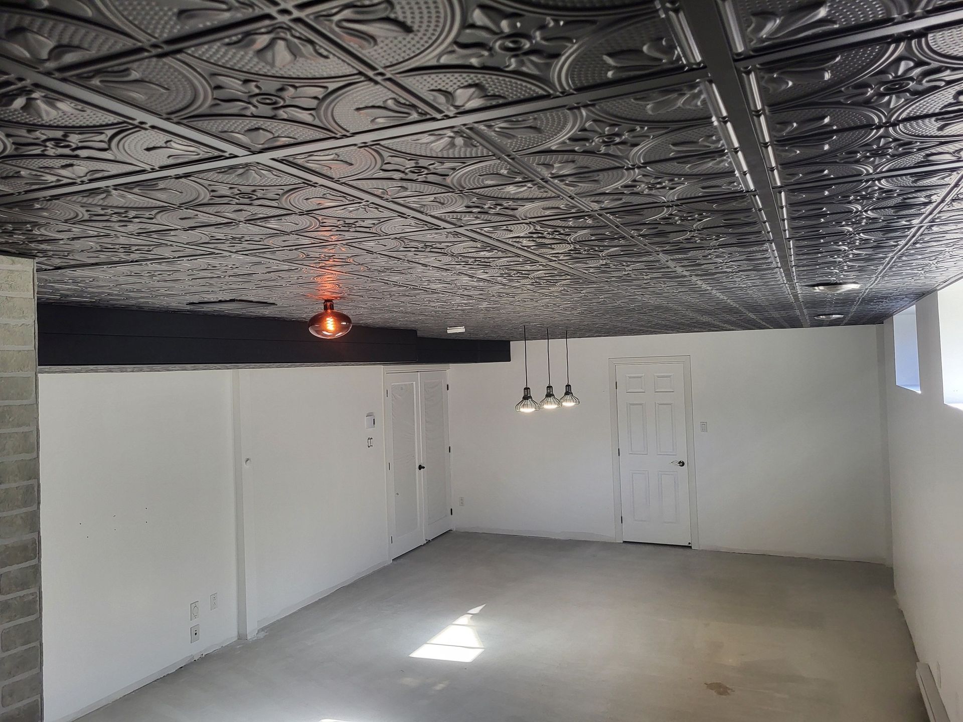Sous-sol intérieur avec plafond en carrelage foncé orné, murs blancs, sol en béton et plusieurs portes. Un luminaire est suspendu au plafond.