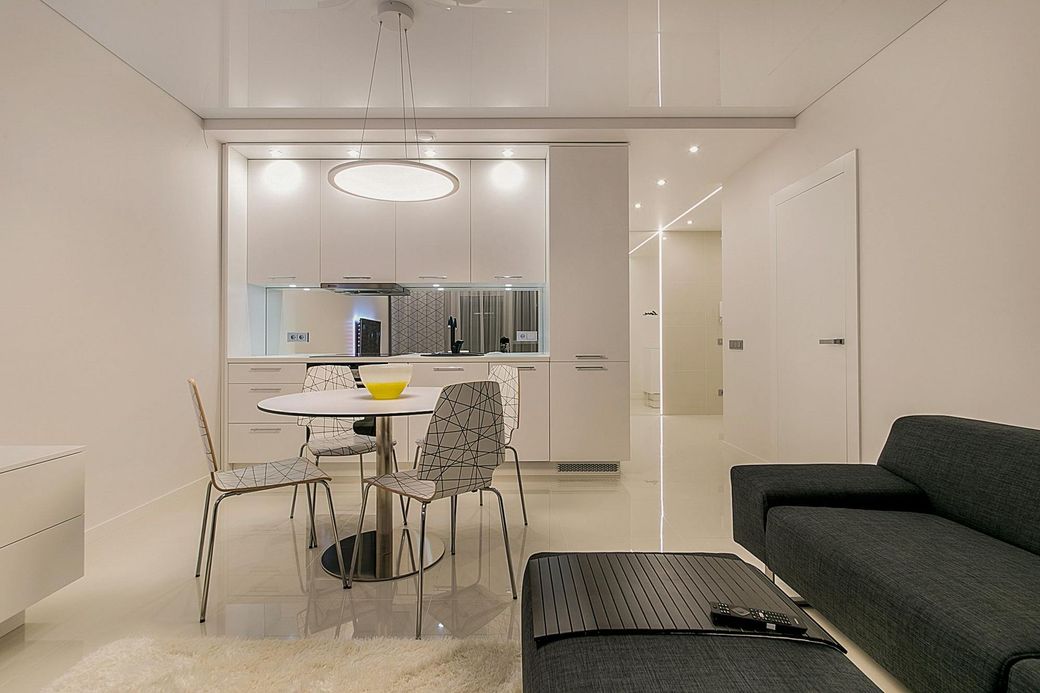 Appartement moderne, entièrement blanc, avec cuisine, salle à manger et salon. L'intérieur comprend une table ronde, des chaises en métal et un canapé gris foncé.