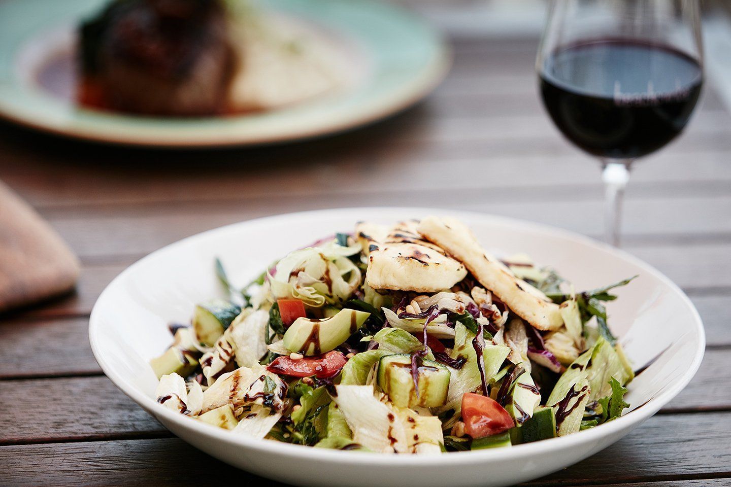 A bowl of salad is on a wooden table next to a glass of wine.