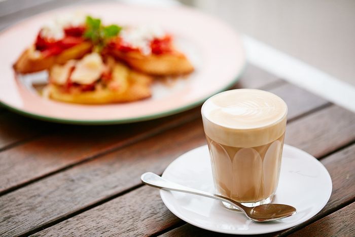 A cup of coffee on a saucer with a spoon and a plate of food in the background.