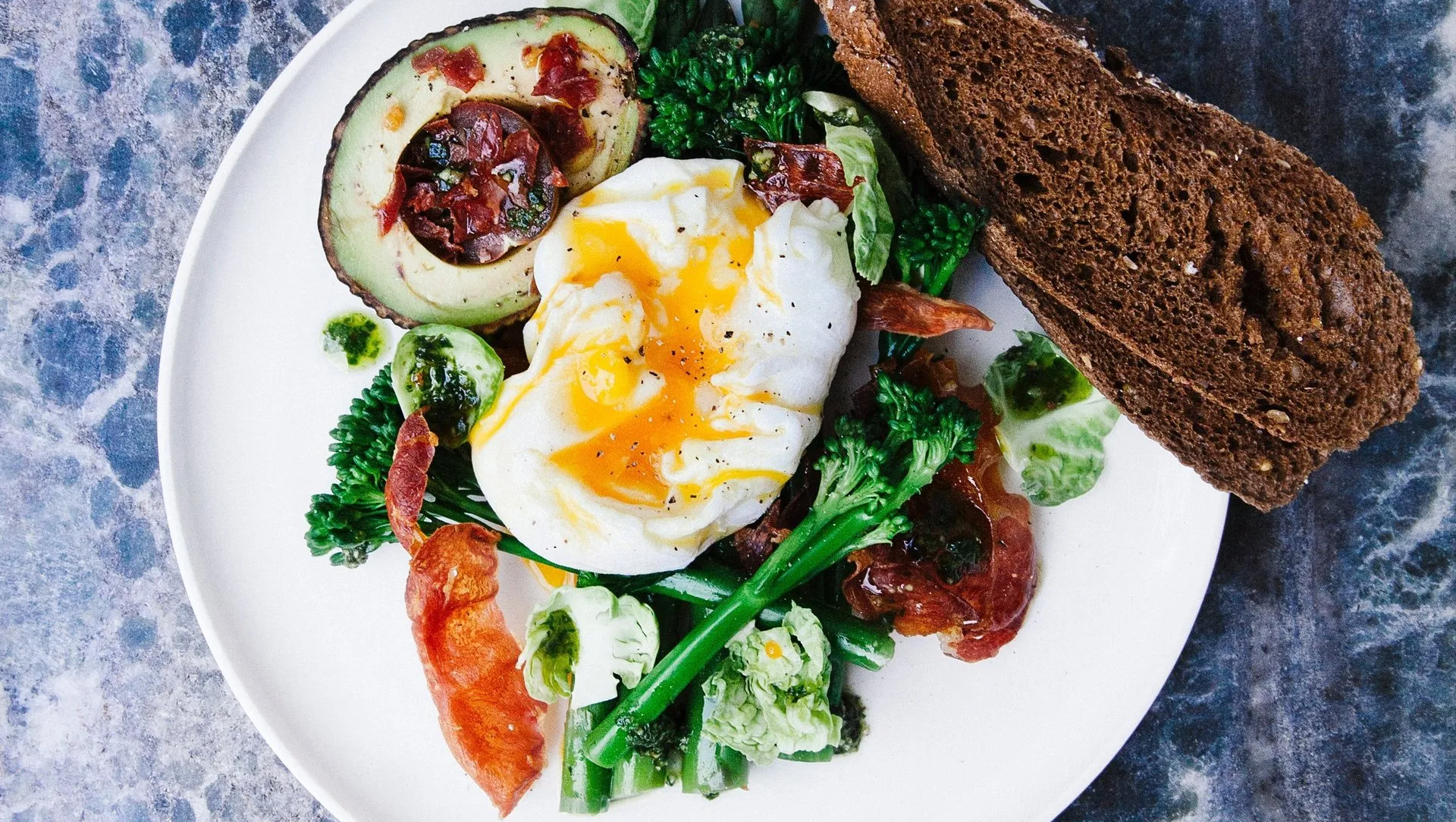 A plate of food with eggs , avocado , broccoli , bacon and bread on a table.