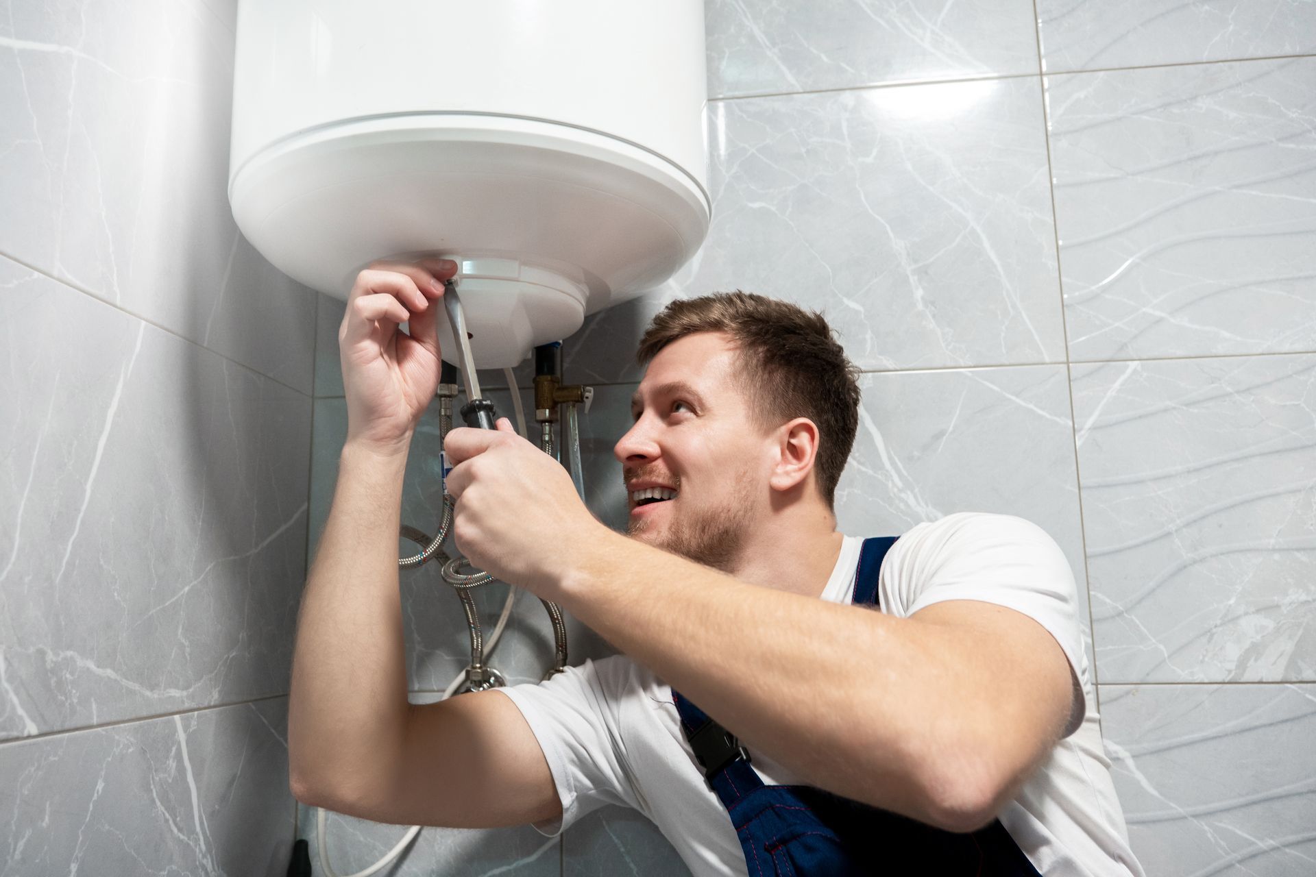 Man repairing a white wall-mounted water heater in a tiled bathroom
