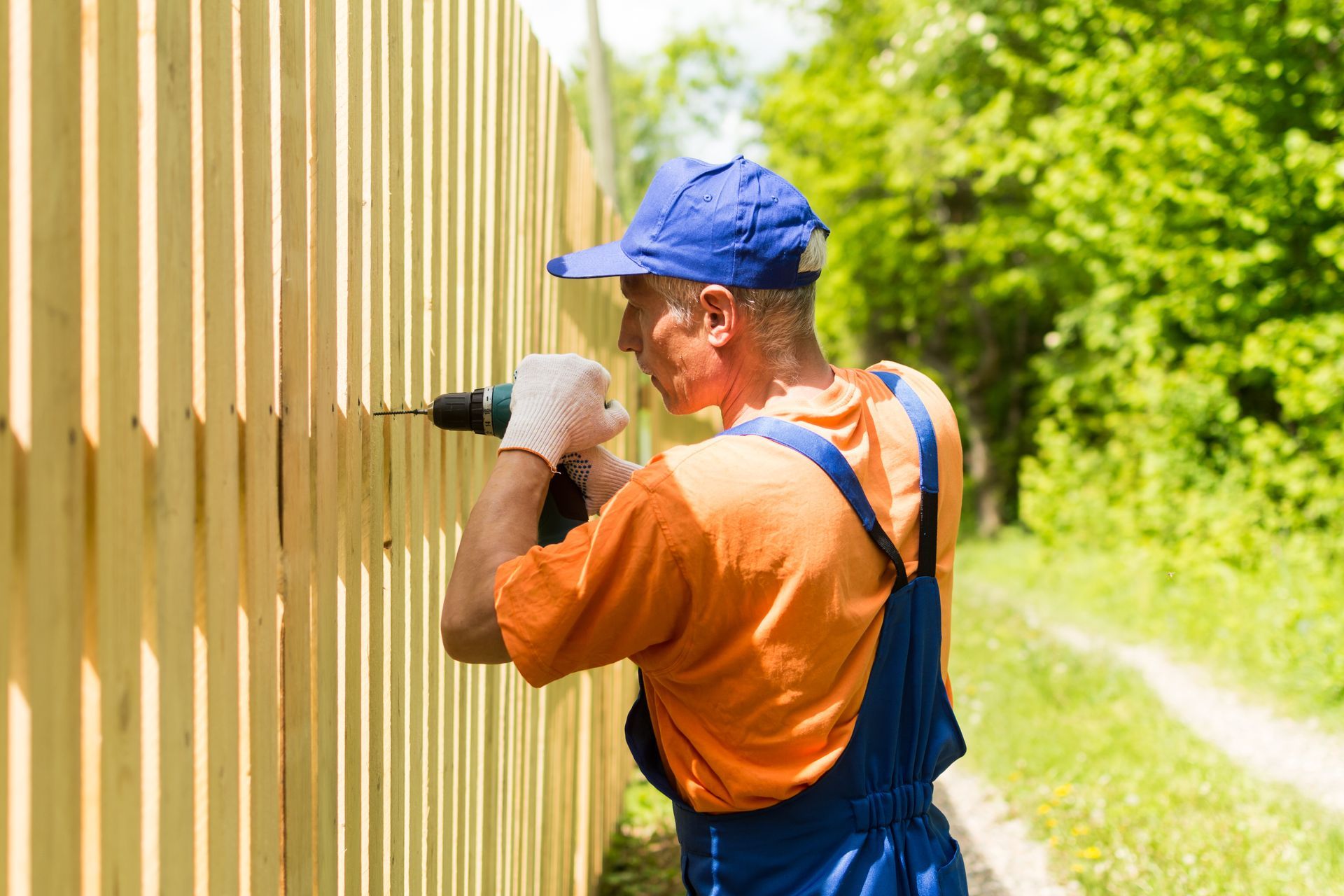Worker in blue overalls and orange shirt drilling into a wooden fence outdoors.